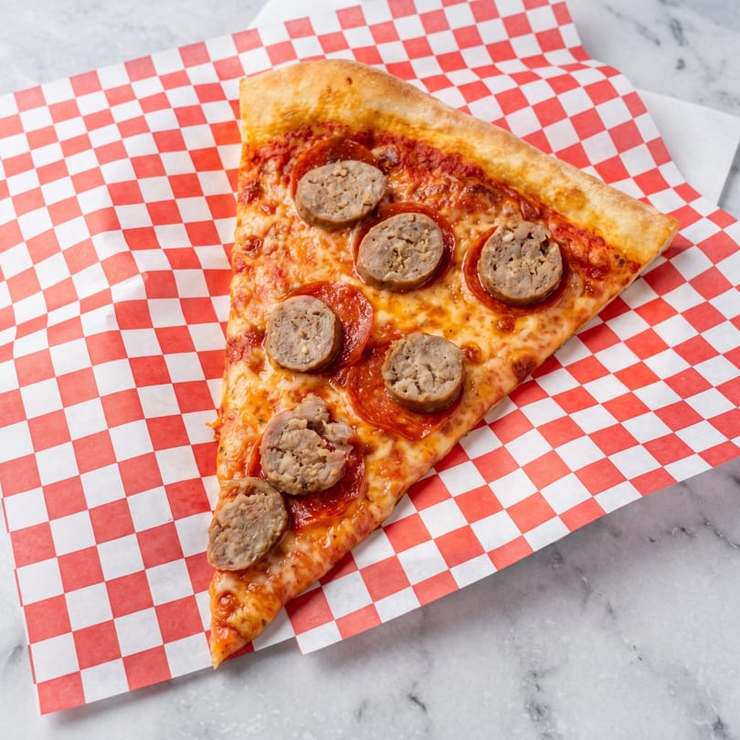 A slice of pepperoni and sausage pizza on red and white checkered paper.