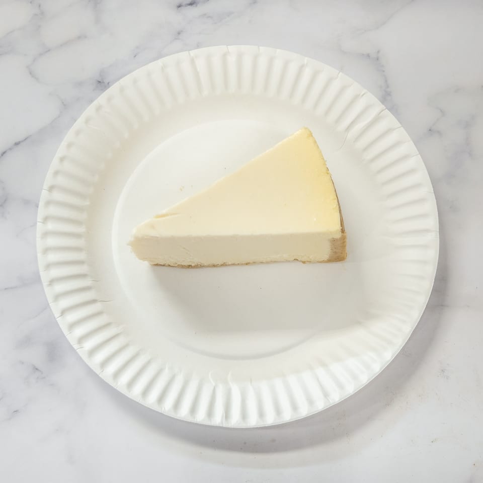 A single slice of classic cheesecake on a white paper plate.