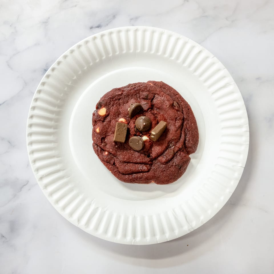 Red velvet cookie with chocolate chips and candy bar pieces on a white paper plate.