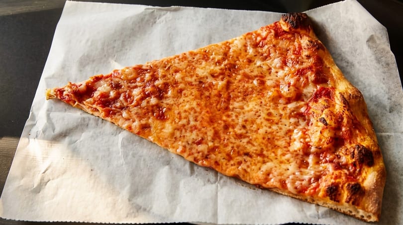 A single slice of cheese pizza with a crispy crust rests on white parchment paper.