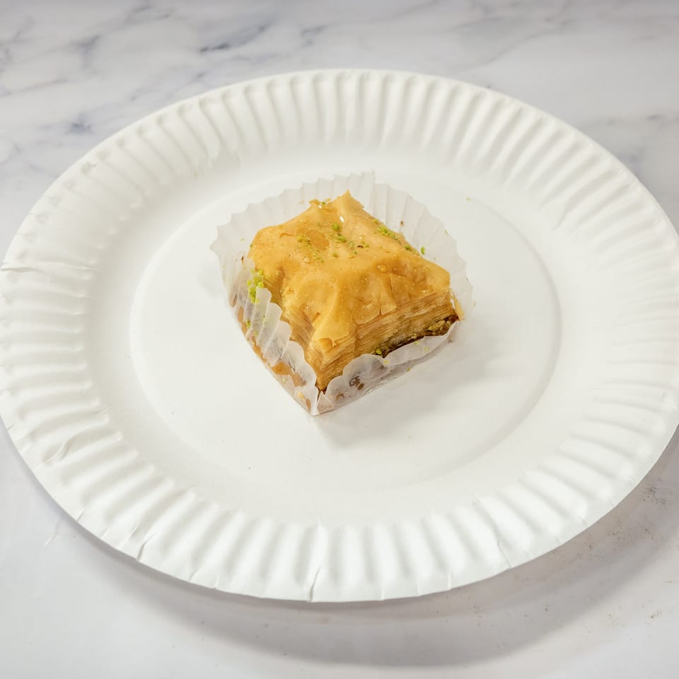 Golden-brown baklava with pistachios in a paper liner on a white paper plate.
