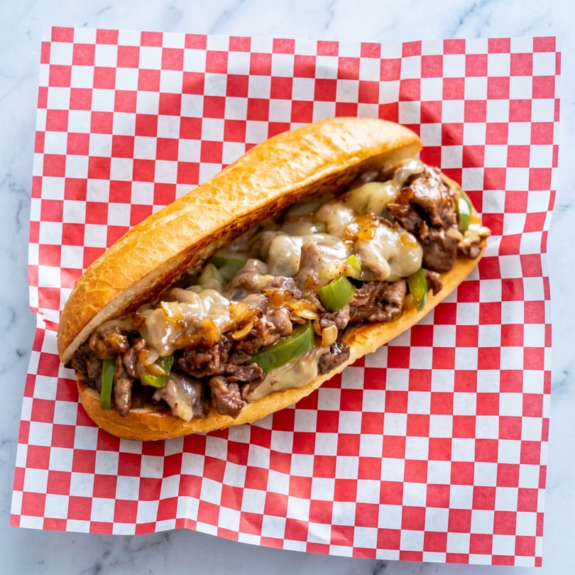 Cheesesteak sandwich with steak, melted cheese, green peppers, and onions on a red and white checkered paper.