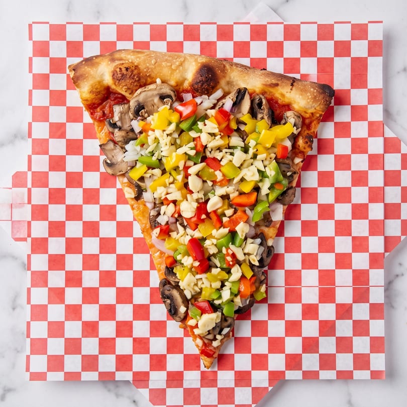 A slice of pizza with mushrooms, onions, bell peppers, and garlic on red and white checkered paper.